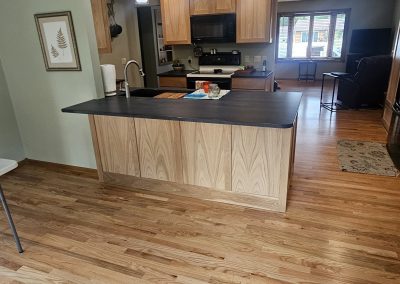 Open kitchen with light wood cabinets, black countertop, three pendant lights, stainless steel appliances, and hardwood floors. The kitchen opens to a living area with large windows and a TV. Decorative wall art above.