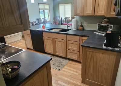 A modern kitchen with light wood cabinets, black countertops, and wood flooring. Pendant lights hang from the ceiling, and appliances and kitchen items are on the counters. A dining area is visible in the background.