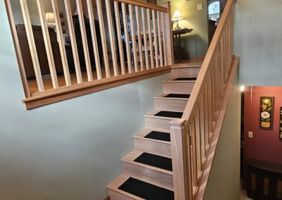 A wooden staircase with beige carpeting leads up to a living area with green walls, a clock, and a lit table lamp. The stairs have black treads and a wooden handrail. A hallway and wall art are visible below.