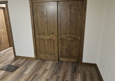 A room with wood-look flooring, cream-colored walls, and a wooden double closet door. An open doorway is visible to the left, showing more of the same flooring. A small dark mat lies on the floor near the doorway.