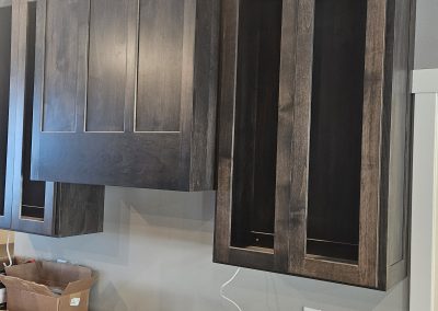 Dark wood kitchen cabinets are being installed above a stove. The cabinet doors are not yet attached, and tools, glue, and screws are spread out on the countertop below. The wall behind is painted light gray.