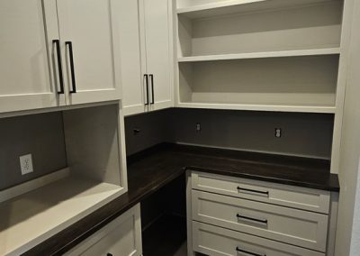Modern walk-in pantry with light gray cabinets, dark countertops, open shelves, drawers, and wood flooring under recessed ceiling lighting. Room is empty and freshly finished.