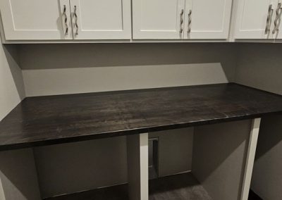 A small room with white cabinets above a dark wooden countertop supported by white legs, and gray wood-style flooring. The walls are light gray and the space appears clean and empty.
