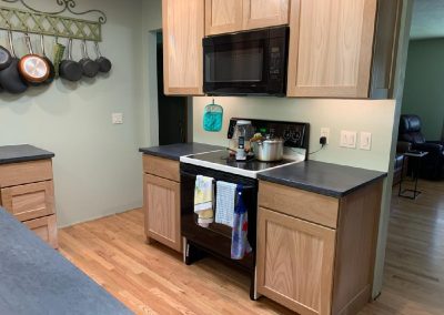 A kitchen with light wood cabinets, black countertops, a black microwave above a stove, pots hanging on the wall, and various kitchen items on the stove. Towels are hanging from the oven door.