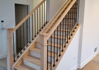 A wooden staircase with black metal balusters and unfinished wood handrails, posts, and steps, in a modern interior space under construction, with protective covering on the floor.