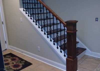A staircase with dark wood steps, a wooden handrail, and black decorative iron balusters sits in a tiled entryway. A floral rug is on the floor near the base of the stairs.
