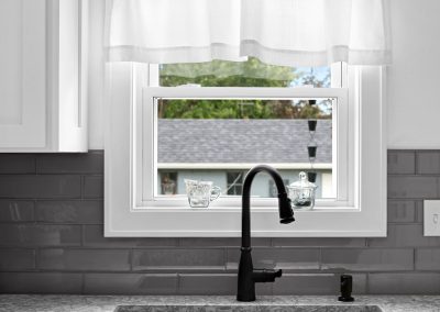 Modern kitchen sink with a black faucet under a window. Light gray countertop, dual basin, gray tile backsplash, white cabinets, and a sheer white curtain. Two glass items sit on the window ledge outside.